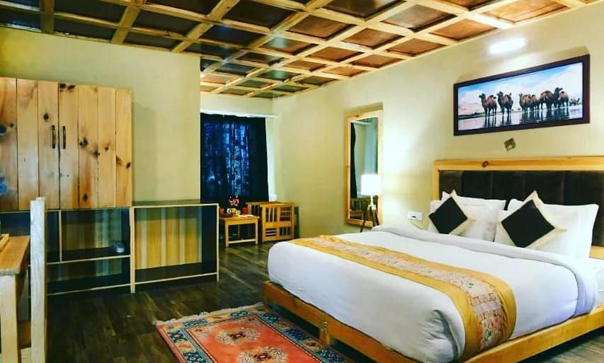 Deluxe Cottage viewed from a corner angle, highlighting the king size bed with white linens and decorative pillows, a patterned rug, and a unique ceiling design at White Sands Resort, Ladakh.