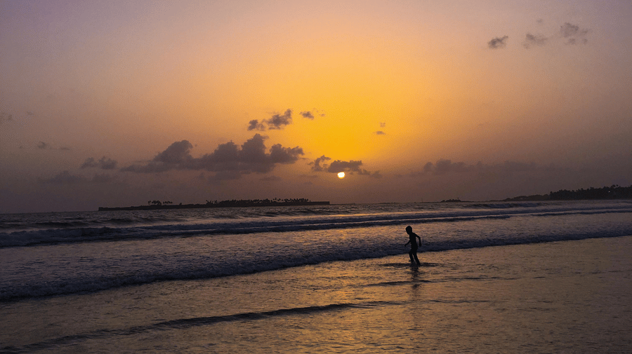 A person stands in the surf at dusk as the sun sets over the ocean, casting a golden glow across the purple sky. Aksa Beach is one of the best sunset points in Mumbai.