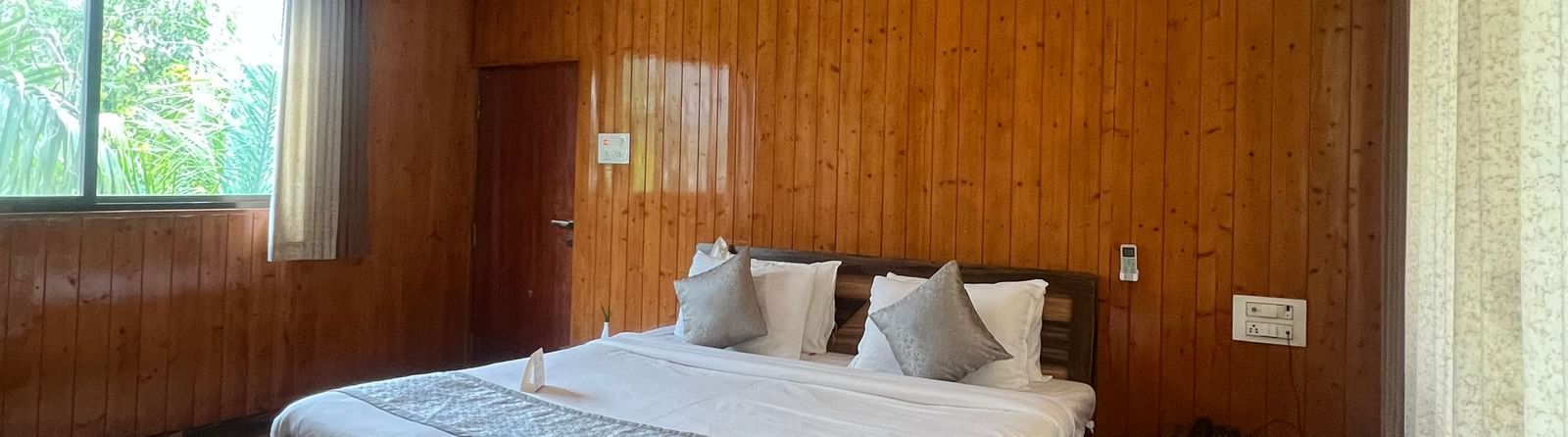 Wide view of the Daksh Elite Cottage, highlighting its wooden interiors, a window with curtains opened, and its centrally placed double bed, at Daksh The Valley Resort, Saputara.