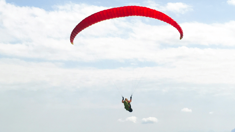Paraglider descending beneath a red canopy over forested mountain slopes, with distant blue ridges fading into the hazy horizon away