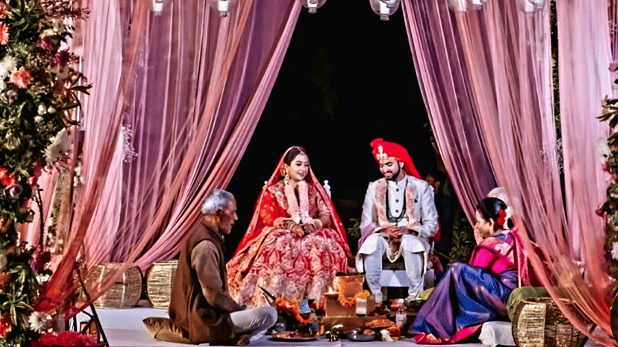 A bride and groom seated under a beautifully decorated wedding mandap at Vedic Village Spa Resort featuring pink drapes floral arrangements and lush green foliage surrounding the couple.