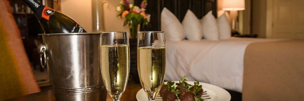 Front view of two champagne glasses filled with champagne , a steel ice bucket and a plate of food on a table in dimly lit room with a neatly made bed and lamps on either side of the bed
