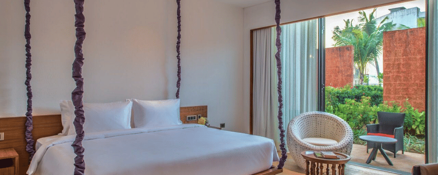 The Deluxe Patio King Room with a large wooden bed hanging from the ceiling on purple rope vines, and an armchair at Azaya Beach Resort, Goa.
