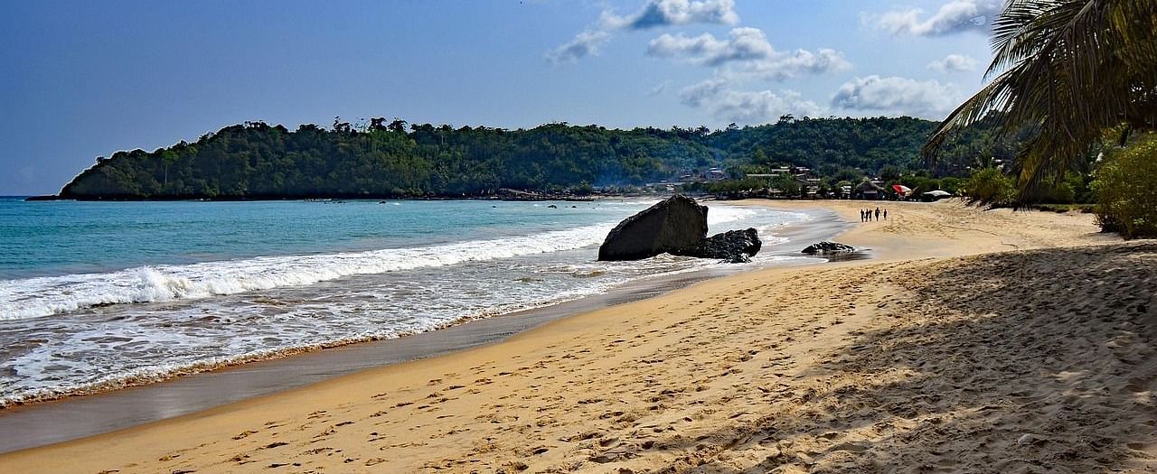 A tranquil view of the Beach with golden sand, turquoise waves, palm trees, and a rocky shoreline with a lush green hills at the backdrop.