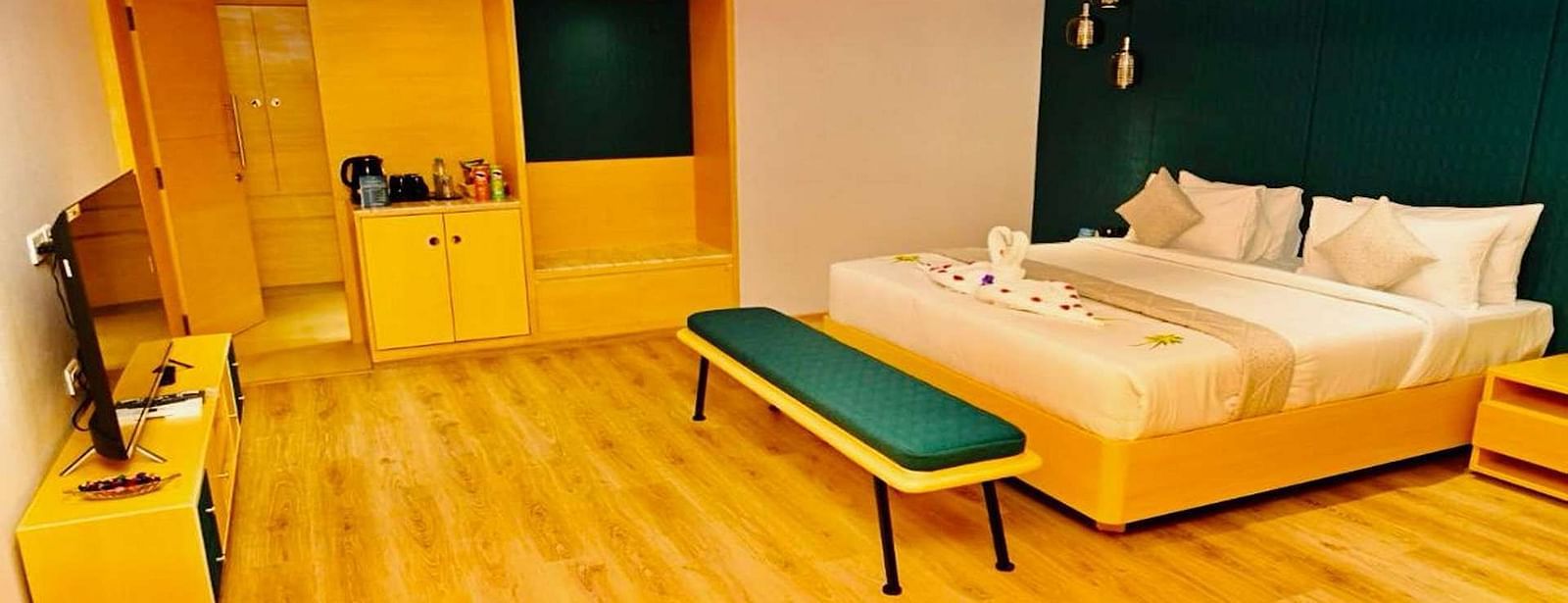 A Presedential Suite featuring a King size bed with white linens and decorative swans, a green accent wall, yellow furniture, and hardwood floors. - Dhanalakshmi Srinivasan Hotels, Perambalur