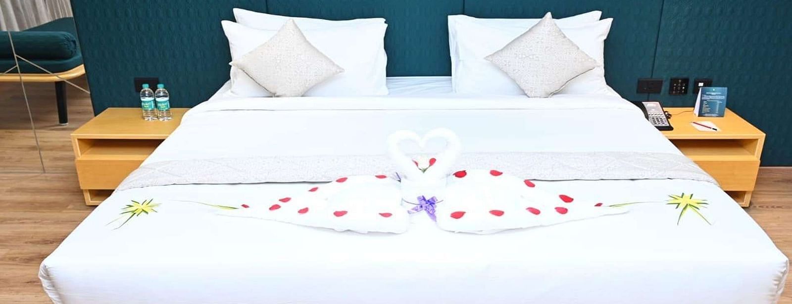 A neatly made Presedential Suite features white linens, two pillows, decorative towel swans forming a heart shape with rose petals, and bedside tables with water bottles - Dhanalakshmi Srinivasan Hotels, Perambalur