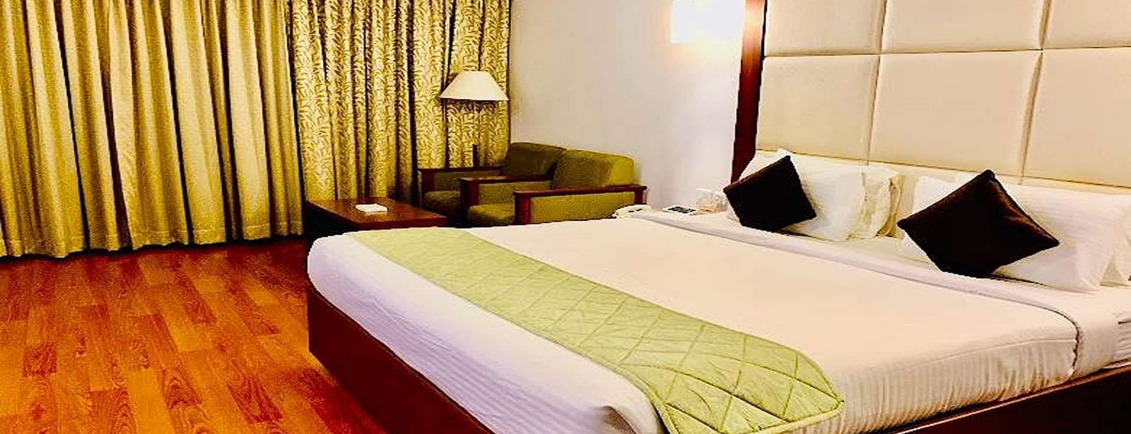 Side view of a Executive Suite at Dhanalakshmi Srinivasan Hotels, Perambalur featuring a bed with white linens, light wood flooring, and a mini-fridge.
