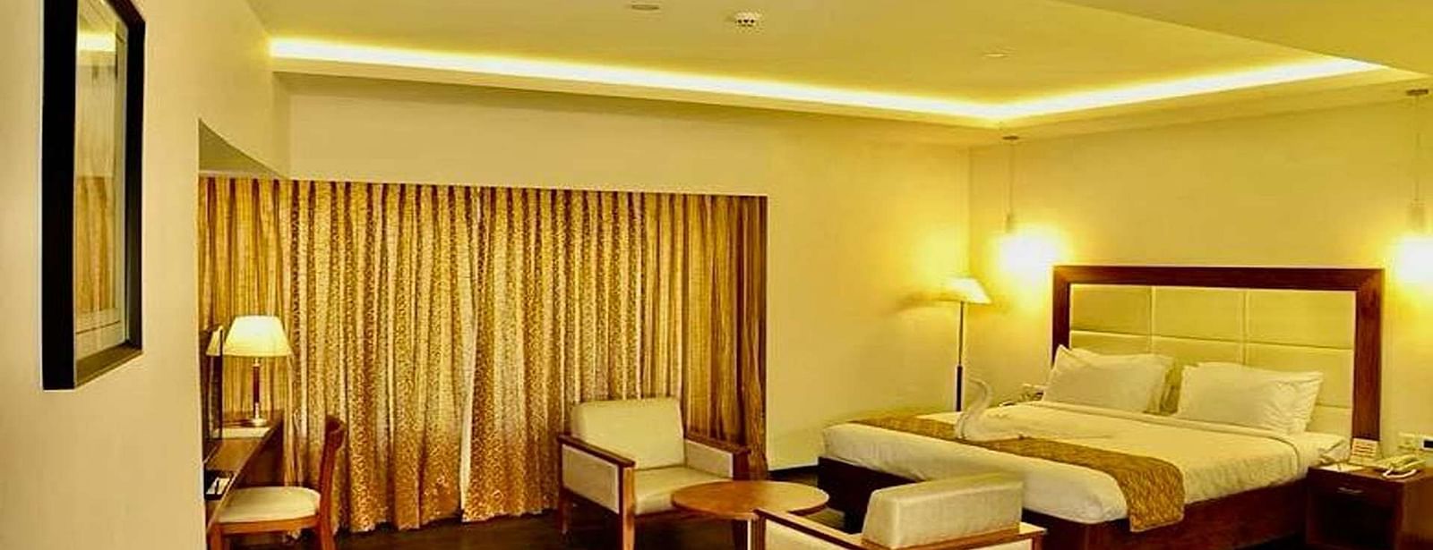 A well-lit Suite at Dhanalakshmi Srinivasan Hotels, Perambalur featuring a king size bed, two armchairs, a small table, and a work desk with a chair.
