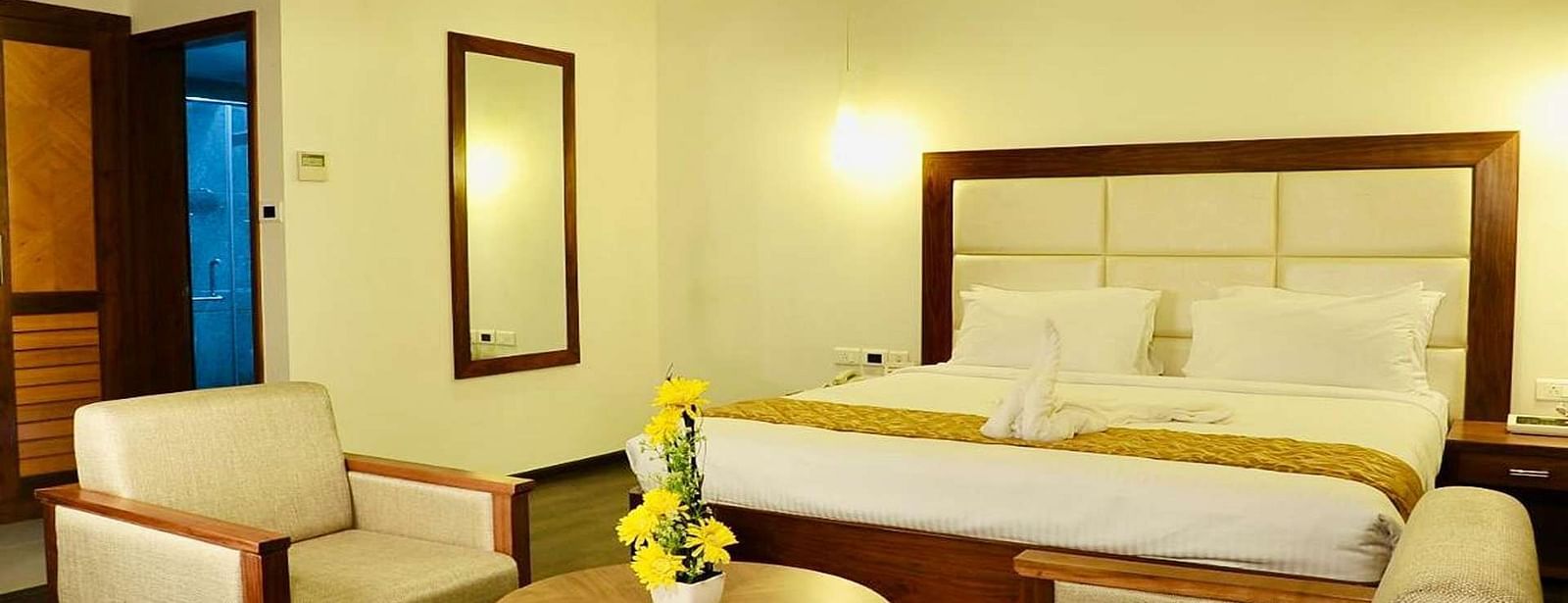 A close up view of well-lit Suite at Dhanalakshmi Srinivasan Hotels, Perambalur featuring a king size bed, two armchairs, a small table, and a work desk with a chair.