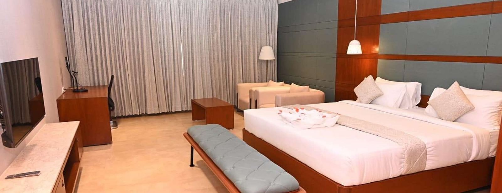 A Executive Suite at Dhanalakshmi Srinivasan Hotels, Perambalur featuring a neatly made bed with white linens and pillows, a wooden headboard, and a desk with a chair.