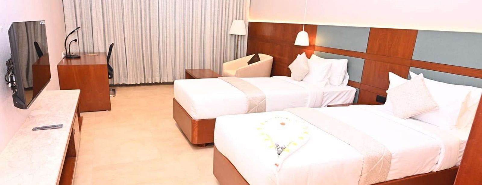 A Executive Suite at Dhanalakshmi Srinivasan Hotels, Perambalur featuring a neatly made twin beds with white linens and pillows, a wooden headboard, and a desk with a chair.