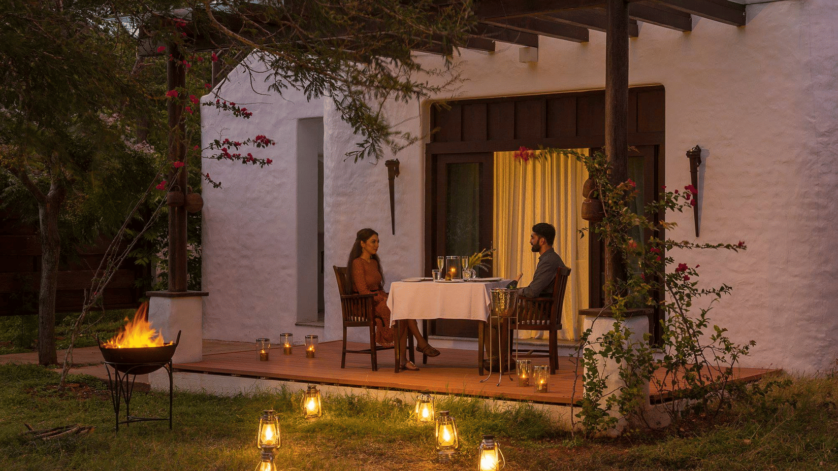 A couple enjoying a romantic outdoor dinner on a wooden patio at The Serai Chikmagalur. The table is set with glasses and a candle, surrounded by small lanterns on the ground and a fire pit nearby.