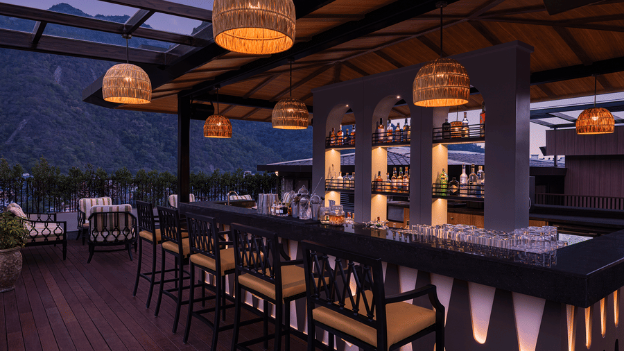 Contemporary bar at The Terrace, Aloha on the Ganges, Rishikesh, featuring high bar stools, illuminated shelving, and panoramic mountain views through glass walls.