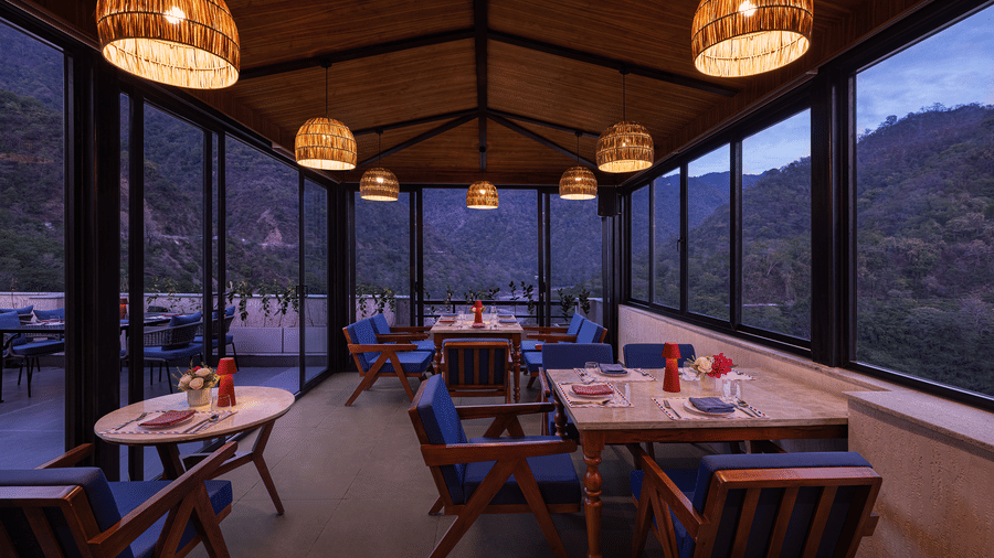Indoor dining area at The Terrace, Aloha on the Ganges, Rishikesh, featuring wooden tables, pendant lighting, and floor to ceiling glass windows overlooking the hills.
