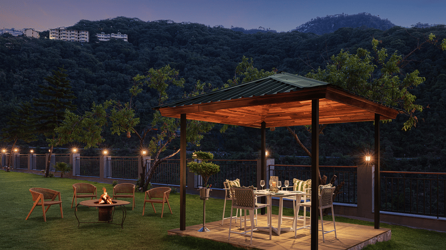 Evening view of an outdoor resort garden with an illuminated dining gazebo and a fire pit area against a dark mountain backdrop.