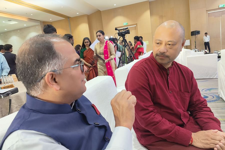 Two men engaged in conversation at a formal indoor event, with one wearing a navy blue vest and glasses, and the other in a maroon kurta.