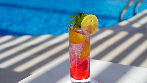a mocktail kept next to the swimming pool with sun's rays hitting the glass - La Maison Hotel, Doha