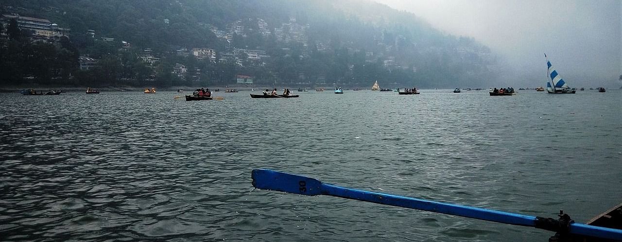 A scenic view from a raft on Nainital Lake, featuring misty hills in the distance and fellow boaters gliding across the shimmering blue water, evoking a sense of tranquility and adventure .