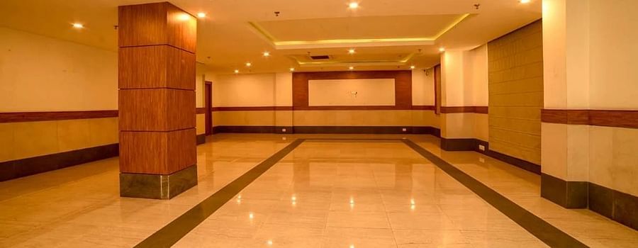 A spacious banquet hall with polished marble flooring, wooden pillars, and a minimalistic design featuring a large open space and a stage at the end.