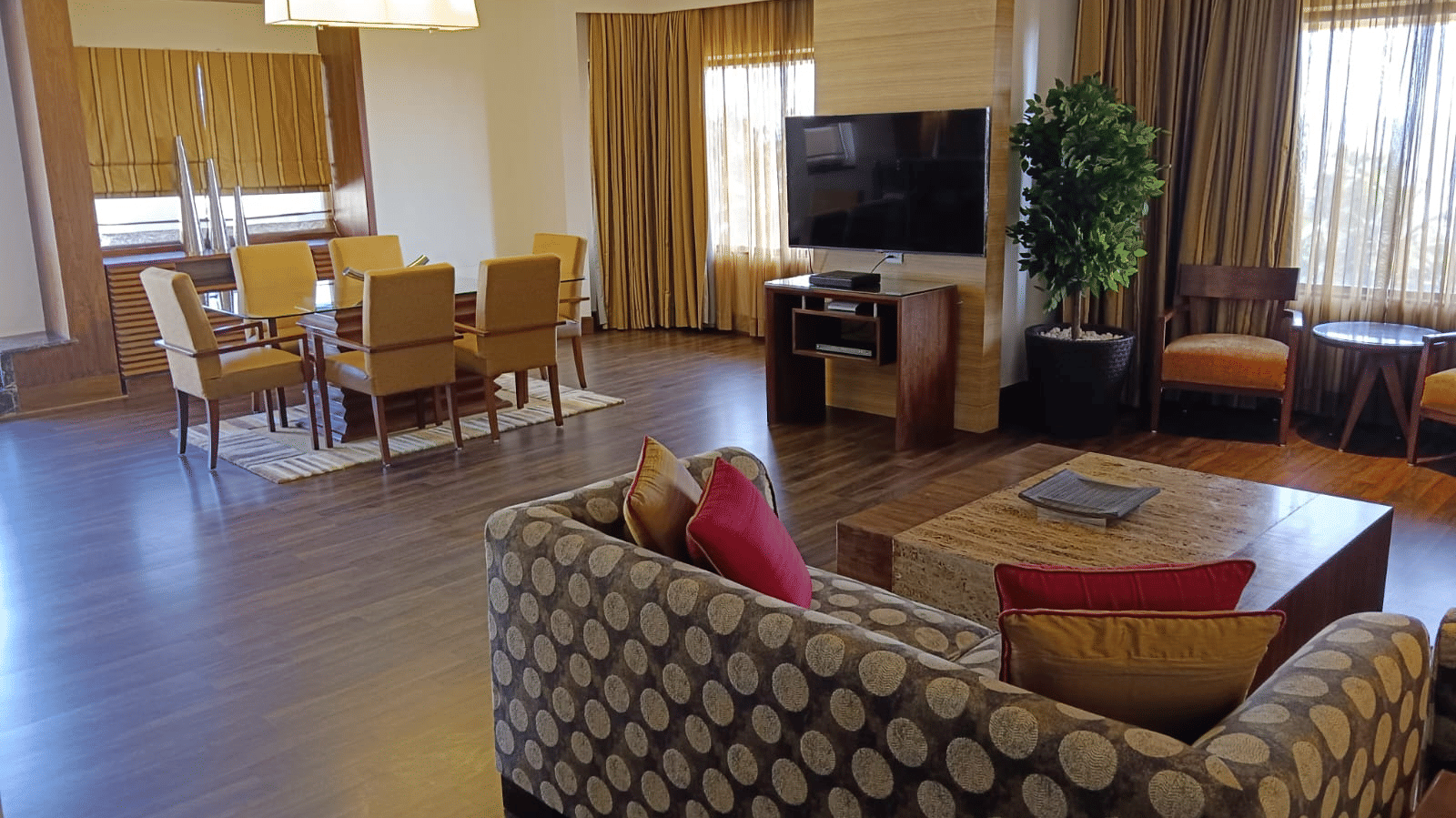Spacious suite living and dining area at Retreat with patterned sofa wooden flooring a wall-mounted TV and a dining table with yellow chairs in a warmly lit open layout.