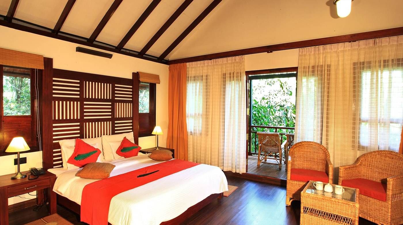 A bed placed in a room with two chairs and table, and a balcony that offers forest views | Vythiri Resort, Wayanad