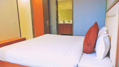 A close-up view of the bed in The Prince Suite at the Prince ZENQ Xpress Resortt Rohtak, featuring a neatly made double bed with white linens, an orange accent pillow, and a doorway open to the bathroom vanity area.
