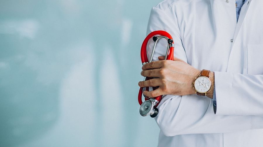 A doctor in a white coat holding a red stethoscope, with arms crossed against a blurred blue background.