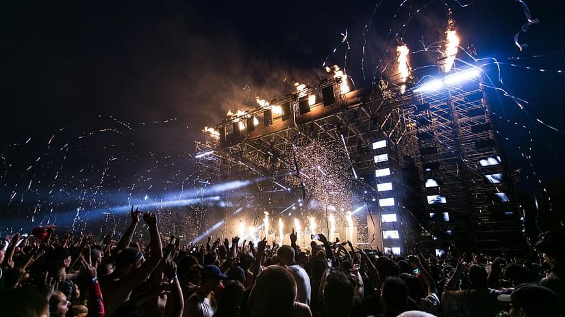 A massive outdoor music festival stage with lights, flames, and confetti before a large, enthusiastic crowd at night.