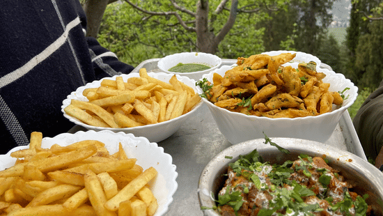 A close-up shot of a tray carrying 2 big servings of french fries, 1 serving of fritters, and 1 serving of an Indian dish garnished with coriander leaves at Amara Upepo - The Sky Village, Manali.