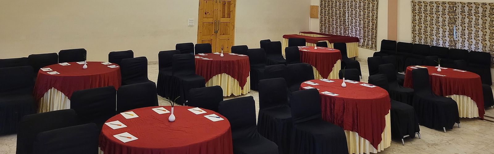 A spacious banquet hall with high wooden ceilings, elegant lighting, and round tables set for events at Dagshai Manor Kasauli - Inde Hotels.
