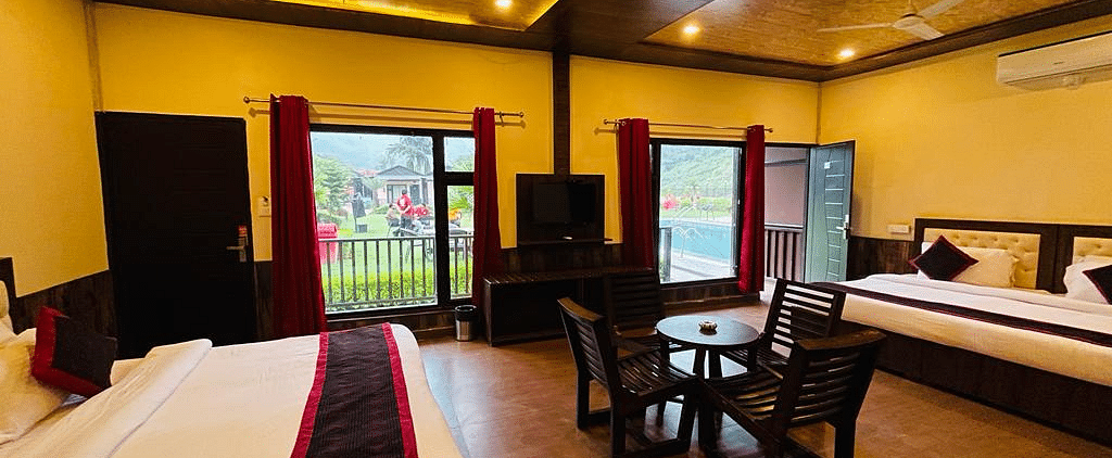 View of the dining table placed in front of a TV with outdoor views on either sides in Family Cottage at DLS MJ River Resort, Rishikesh.