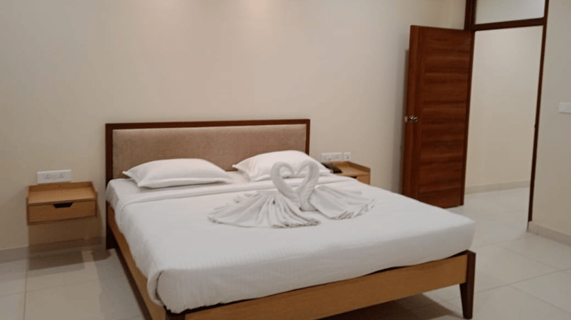 Bedroom at A M Suites with double bed, white bedding, swan-shaped towel sculptures forming heart, beige walls, tiled floor, wooden nightstands, open door, and recessed ceiling lighting.