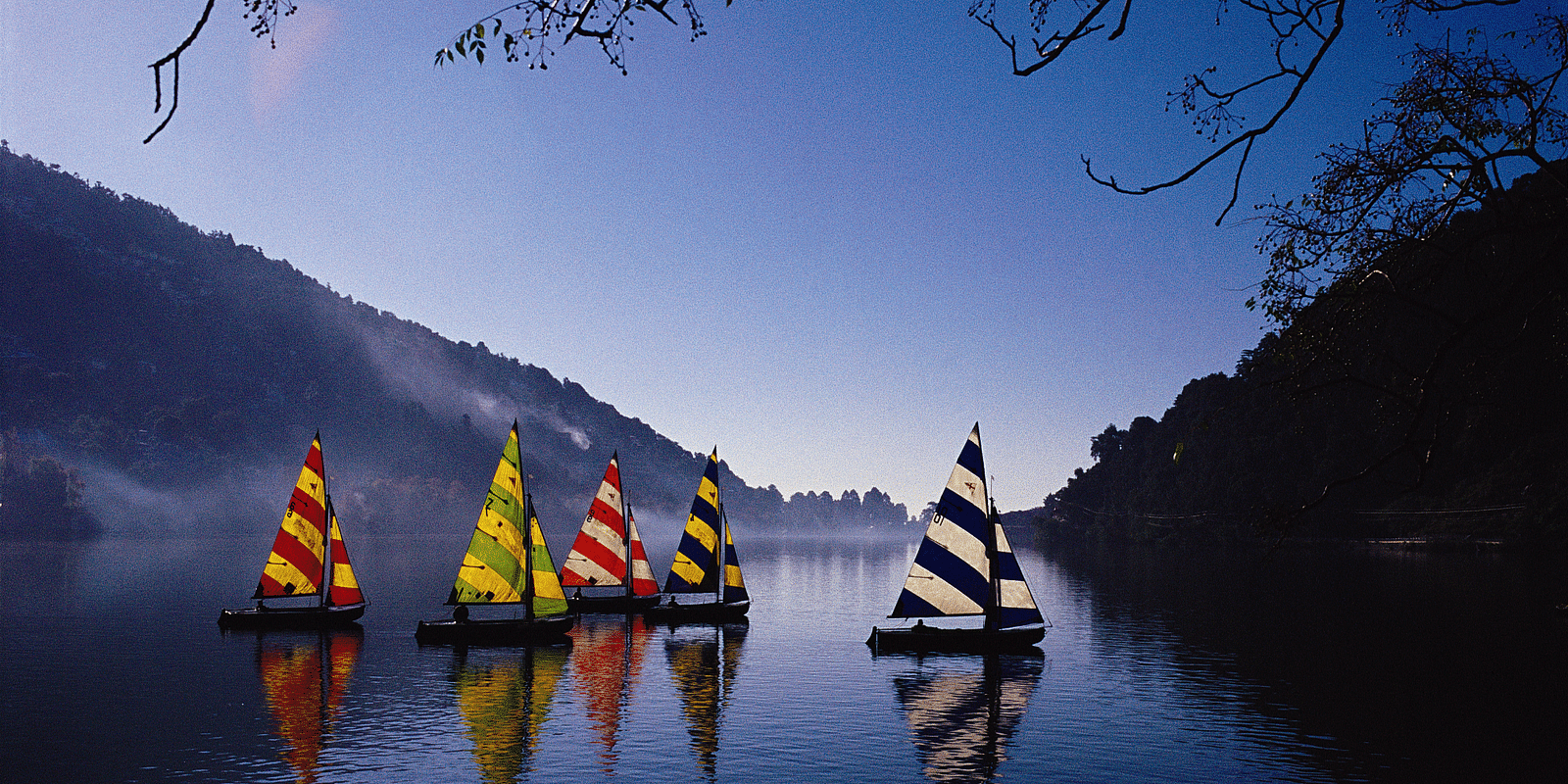 An image with five sailboats pictured on a lake , with their reflections visible on the water, with the magnificent backdrop of the mountains converging.