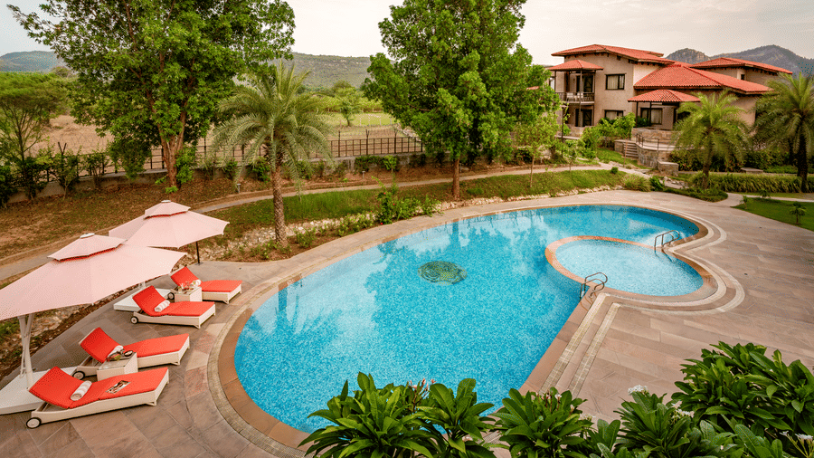 The large outdoor swimming pool area at AamaGhati Wildlife Resort with bright orange sun loungers, umbrella shades, and surrounding resort buildings.