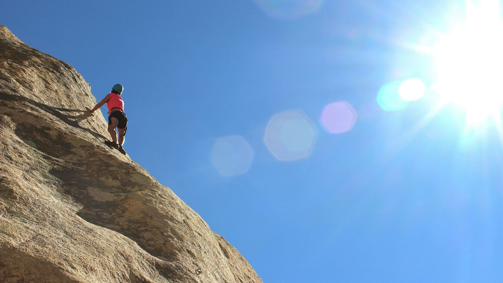 A woman rock climbing a mountain with the sun shining down on her.
