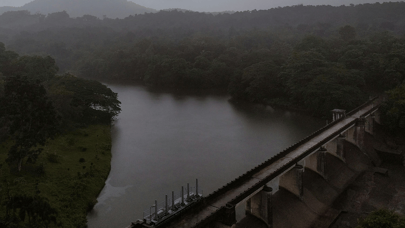 A large concrete dam stretching across a valley between green hills under a cloudy sky.