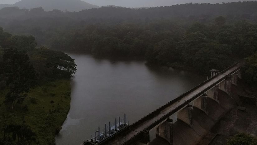 A large concrete dam stretching across a valley between green hills under a cloudy sky.