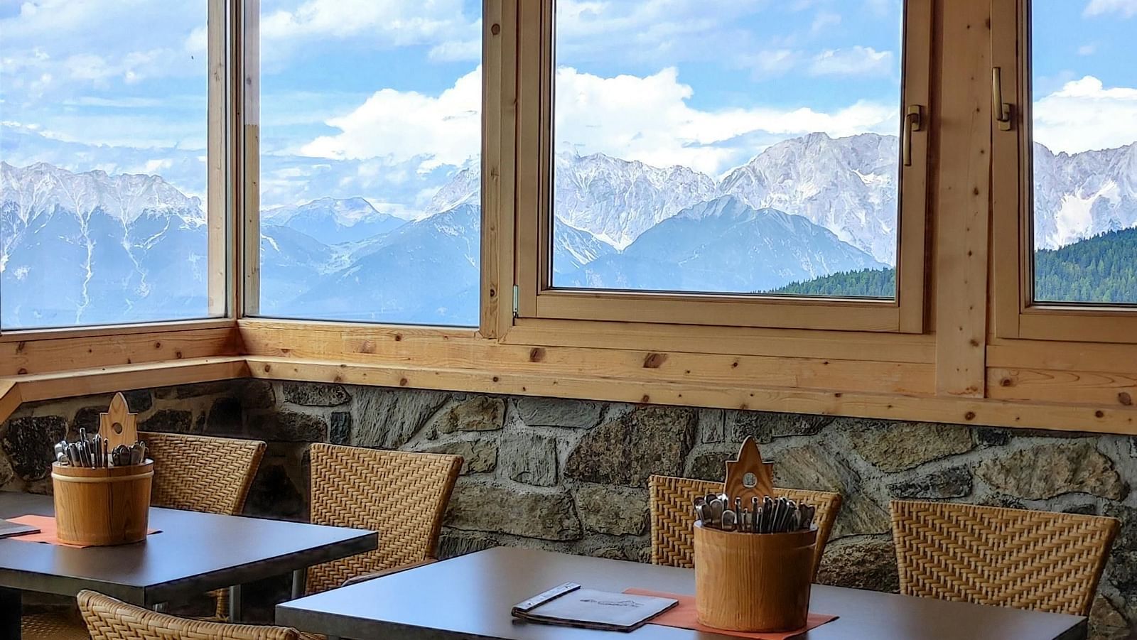 A cosy indoor dining area with wooden ceilings and large windows offering a scenic view of snow-capped mountains under a partly cloudy sky.