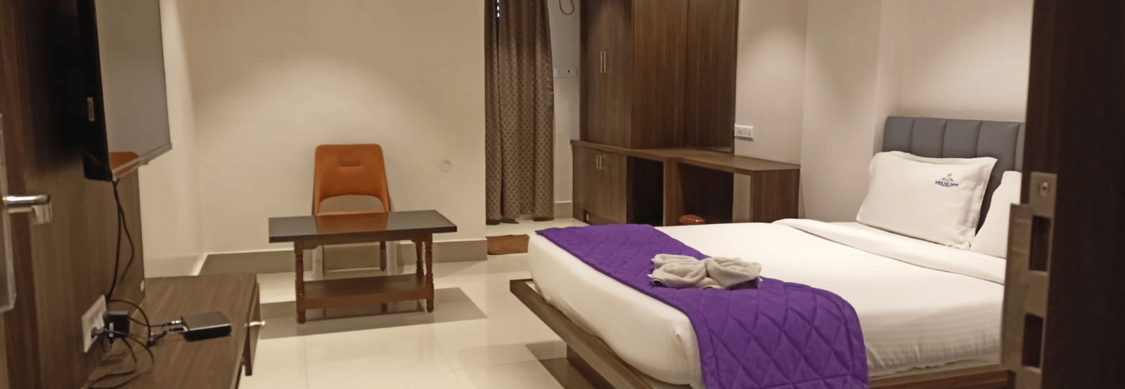 A view of a guest room showing a wall-mounted television, a small wooden desk, and a bed.