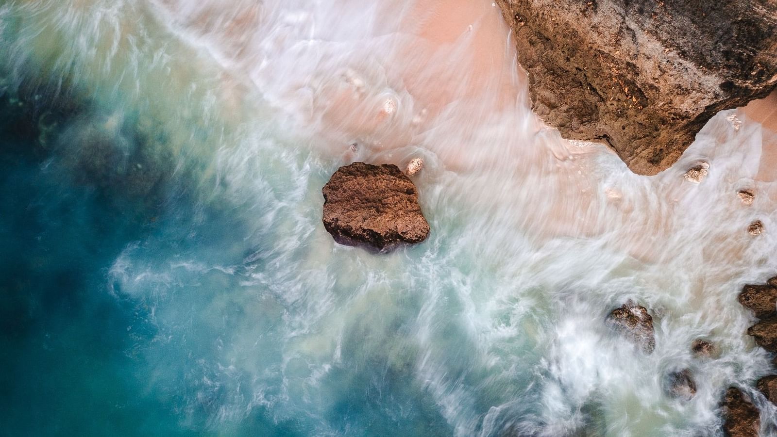 An aerial view of a coastal beach scene with waves crashing around a solitary large rock