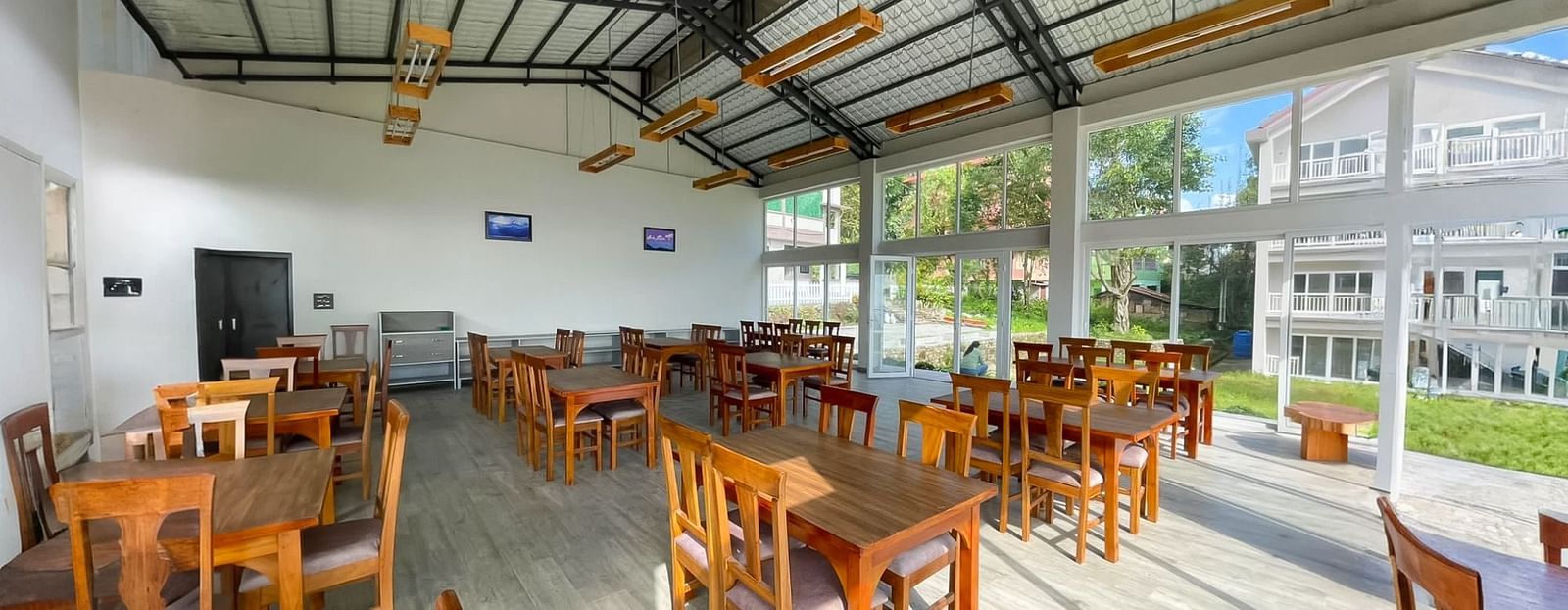 A spacious and bright dining area at Yashshree Resorts & Spa, Kalimpong, featuring numerous wooden tables and chairs, under a high ceiling with exposed beams.