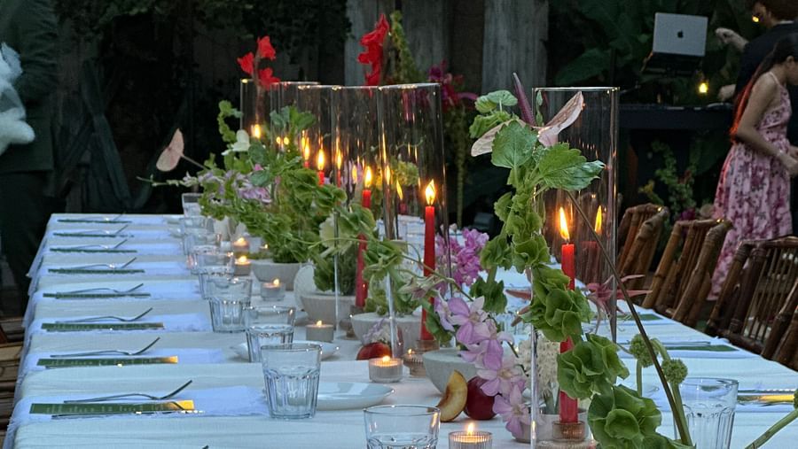 A long dinner table where several items are placed including forks, menu cards, glass,  etc. The table is also decorated with candles wrapped in a glass and artificial flower pots with flowers and leaves.