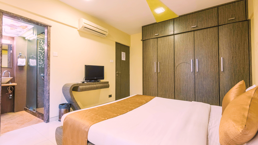 Modern deluxe-room with bed in white sheets and brown pillows, wall?mounted television above shelf, large dark wooden wardrobe, warm lighting, and doorway leading to bathroom with sink and mirror - Dragonfly Hotel - The Art Hotel