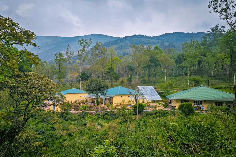 Ibex Resorts, Valparai (Mistly) buildings with green roofs located on a hillside, surrounded by trees, plants, pathways, and mountain ranges in the background under a cloudy sky.
