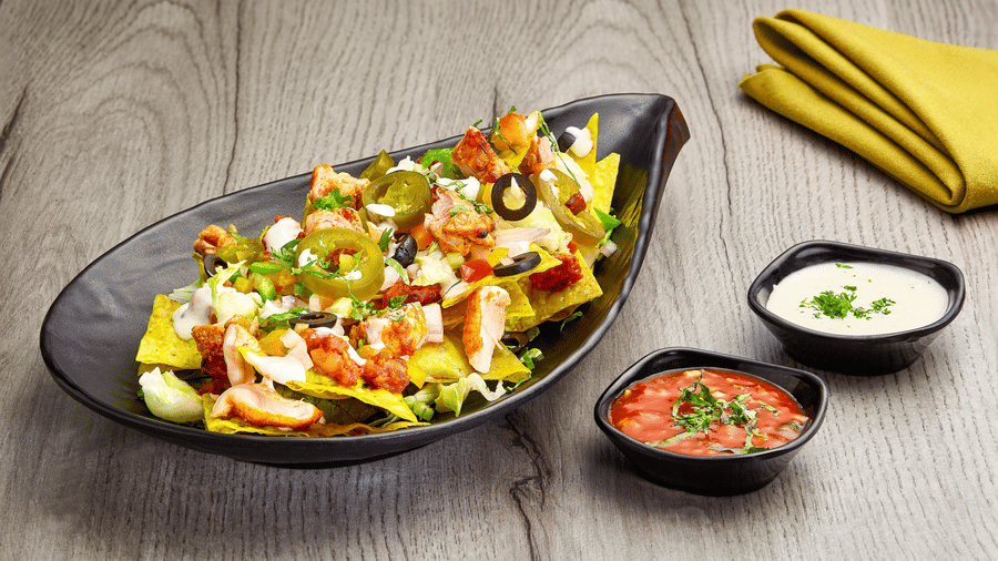 Black boat-shaped platter of chicken nachos with jalapenos, olives, tomato and herbs, served with bowls of salsa and sour cream dip.