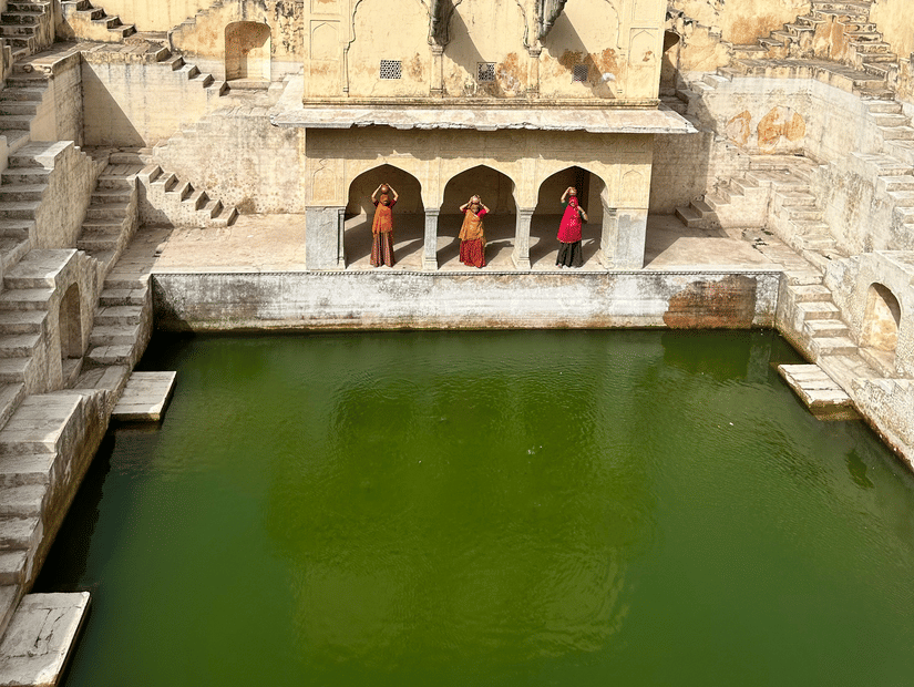 A high-angle view of a stone stepwell containing green water, surrounded by arid hills and local structures.
