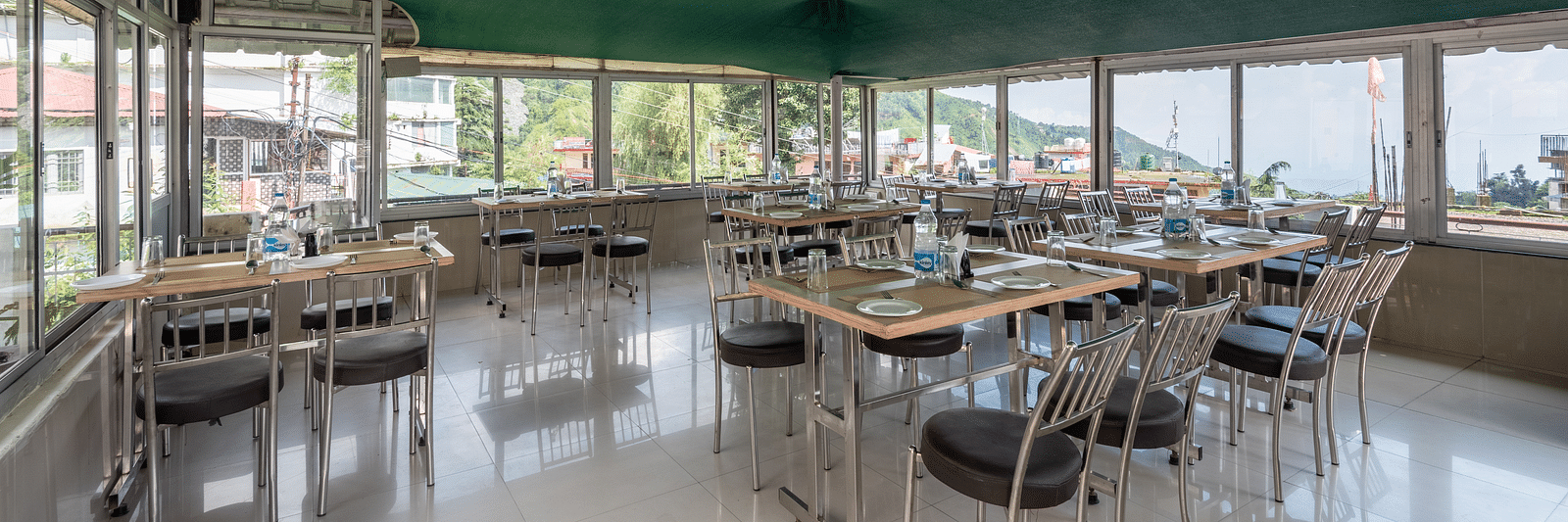 A coffee house with tables and chairs under a green canopy, featuring large windows offering an outdoor view. The tables are set with plates and bottles of water - DLS Meghavan Resort, Dharamshala