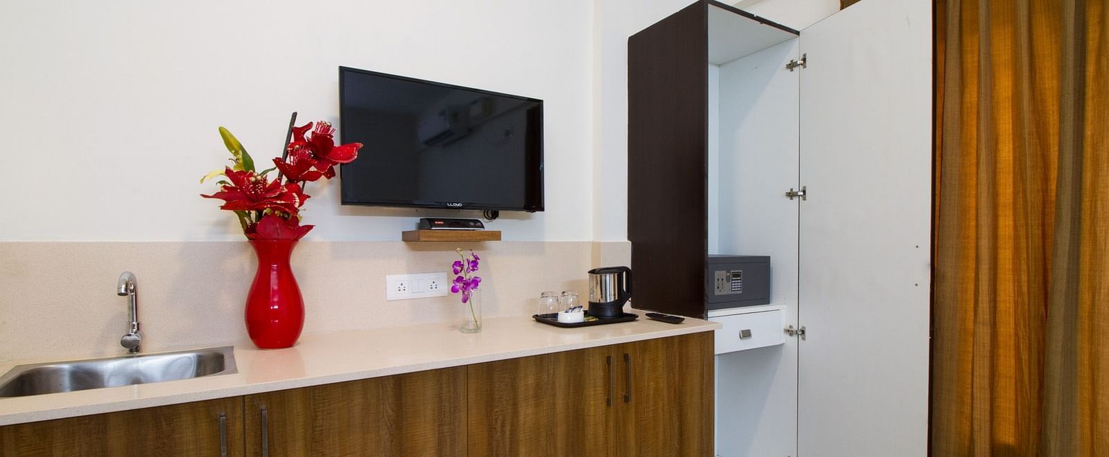 A wall-mounted television and a modern kitchenette area featuring a sink, a red vase, and wooden cabinets at Sanctum Suites Whitefield, Bangalore.