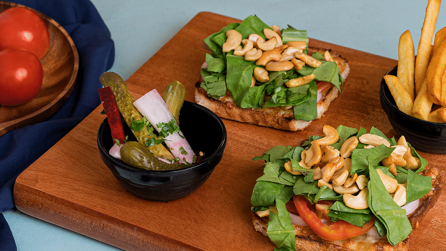 An image of two sandwiches topped with greens and cashews, accompanied by a bowl of French fries and another bowl of vegetable salad.