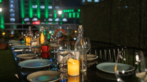 An evening view of a dining table set on a rooftop with artificial grass, candle lighting, and a cityscape backdrop with lit buildings.